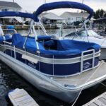 Blue Bennington pontoon boat docked at a marina, with blue canopy and seating, tied to a wooden dock by calm water.
