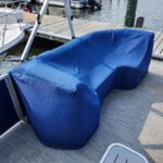 Blue cushioned seating covered with a protective blue cover on a pontoon boat deck, near the dock and water.