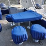 Blue-covered outdoor seating area on a boat deck, with a rectangular table and two round stool seats covered. Water visible in the background.