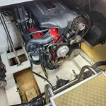 Marine inboard diesel engine with belt-driven pulleys and hoses in a boat engine room; surrounding components and metal deck visible.
