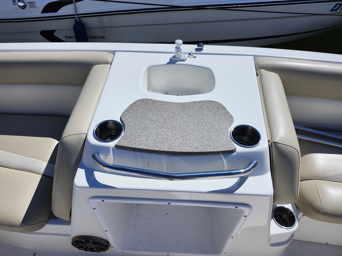 Boat cockpit centerpiece: small sink with faucet, two cup holders, and a textured cover on a white console between beige seats.
