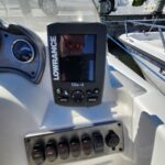 Marine helm with a Lowrance Elite-4 fishfinder mounted on the dashboard and a row of switch controls beneath; boats and a car parked on a docked dockside in the background.