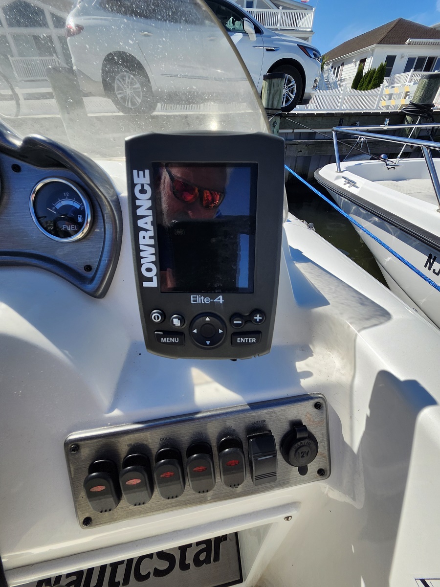 Marine helm with a Lowrance Elite-4 fishfinder mounted on the dashboard and a row of switch controls beneath; boats and a car parked on a docked dockside in the background.