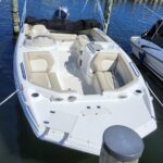 White motorboat moored at a wooden dock, beige cushioned seats and a folded black canopy over the cockpit, calm blue water in the background.