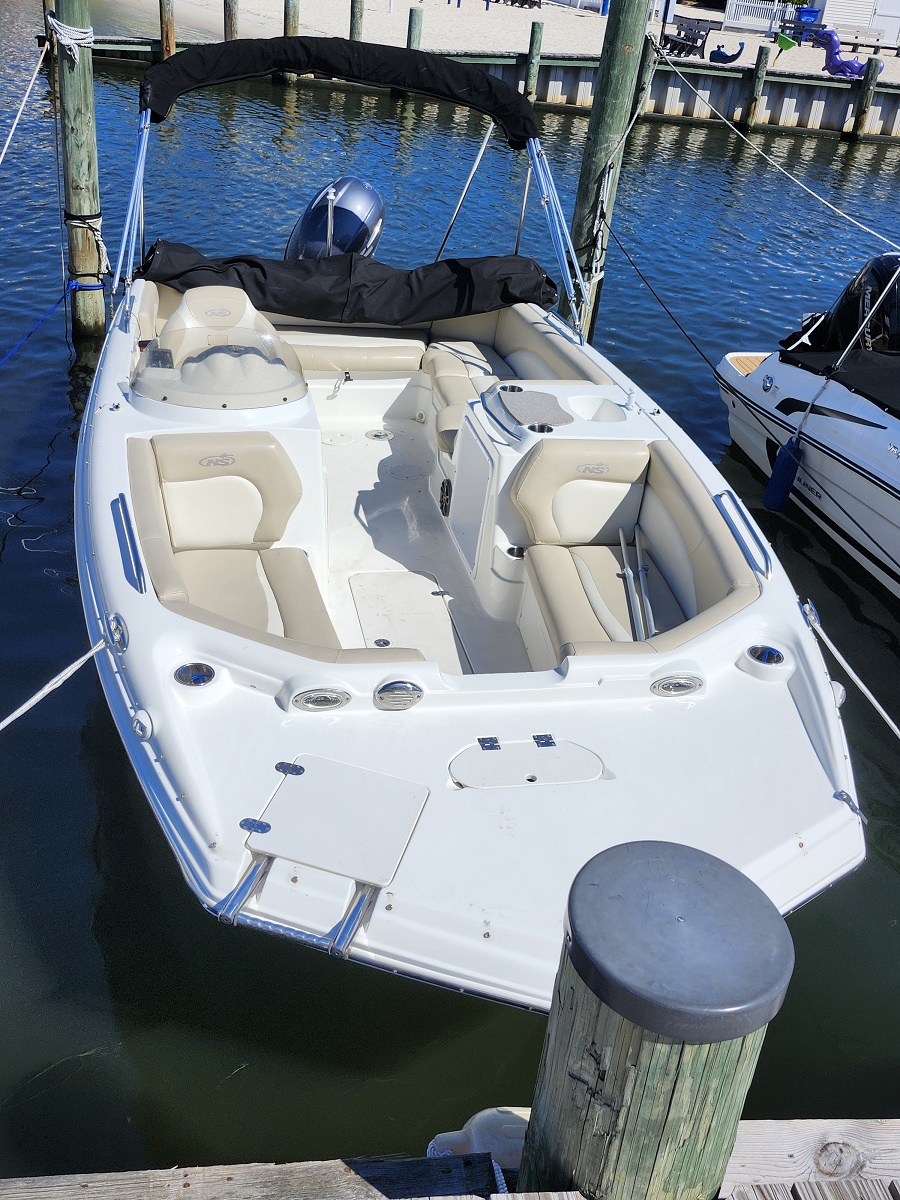White motorboat moored at a wooden dock, beige cushioned seats and a folded black canopy over the cockpit, calm blue water in the background.