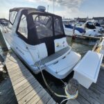 White motorboat docked at a marina with a black canvas canopy and clear side curtains, Larson branding on the side, tied to a wooden dock under a sunny sky.