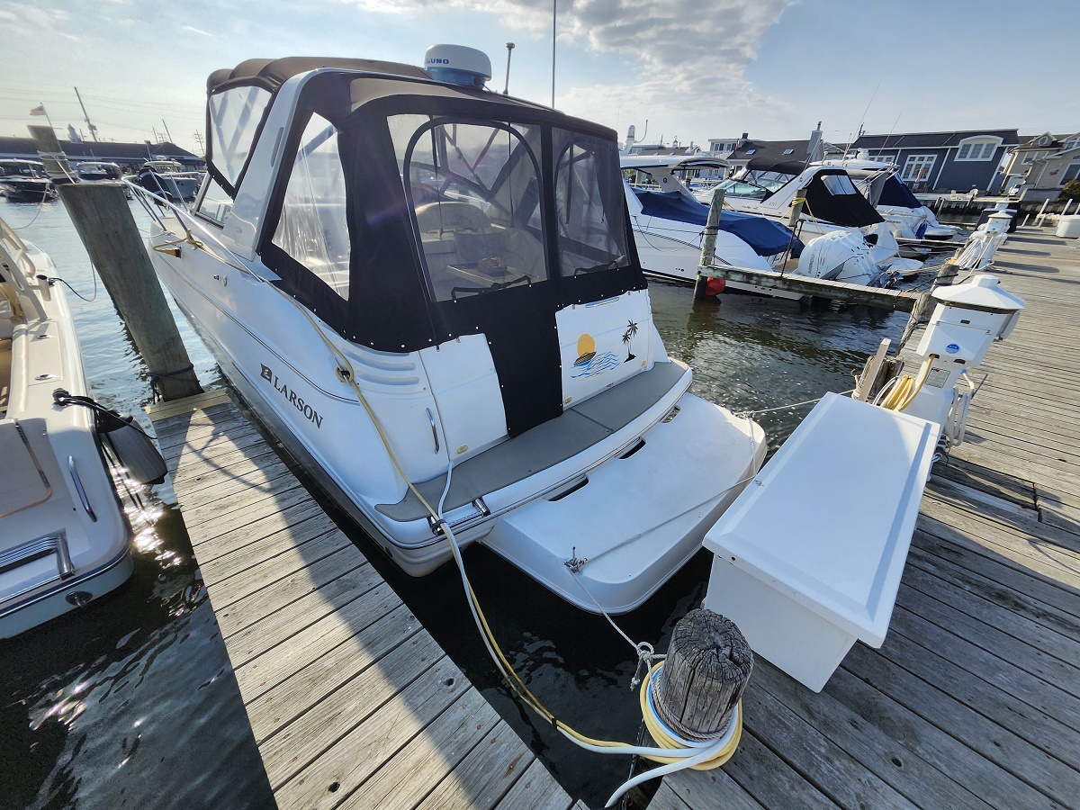 White motorboat docked at a marina with a black canvas canopy and clear side curtains, Larson branding on the side, tied to a wooden dock under a sunny sky.