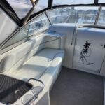 White leather cockpit seating area inside a boat, with a raised black non-slip panel and cup holders; sunlight casts shadows on the cushions near a windowed cabin.