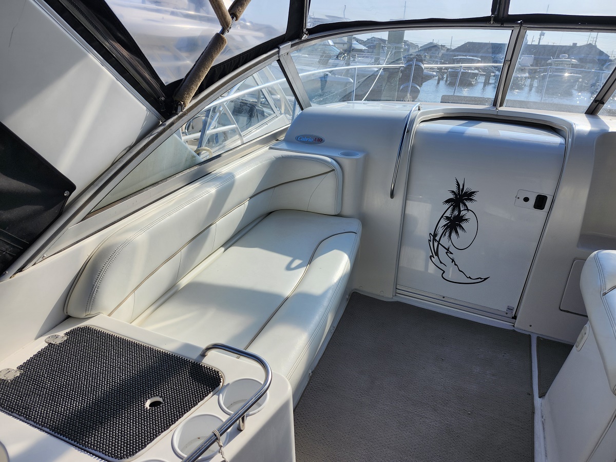 White leather cockpit seating area inside a boat, with a raised black non-slip panel and cup holders; sunlight casts shadows on the cushions near a windowed cabin.