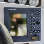 Furuno NAVnet marine navigation display with a control keypad on the right and a perforated steering wheel to the left; gauges in the background