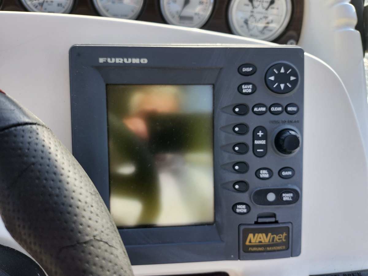 Furuno NAVnet marine navigation display with a control keypad on the right and a perforated steering wheel to the left; gauges in the background