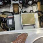 view of boat engine compartment with battery, air filter, hoses and wiring; a person’s boot at the edge of the hatch