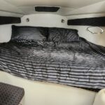 Inside a small boat cabin, a large bed with dark gray striped bedding and two pillows, flanked by circular porthole windows.
