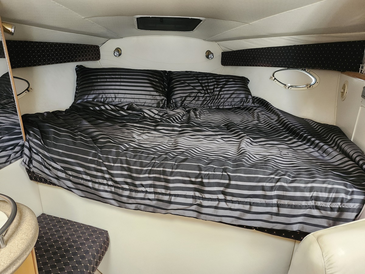 Inside a small boat cabin, a large bed with dark gray striped bedding and two pillows, flanked by circular porthole windows.