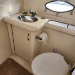Compact boat bathroom with oval porthole window, sink, faucet, soap bottle, toilet, and tissue holder in cream cabinetry