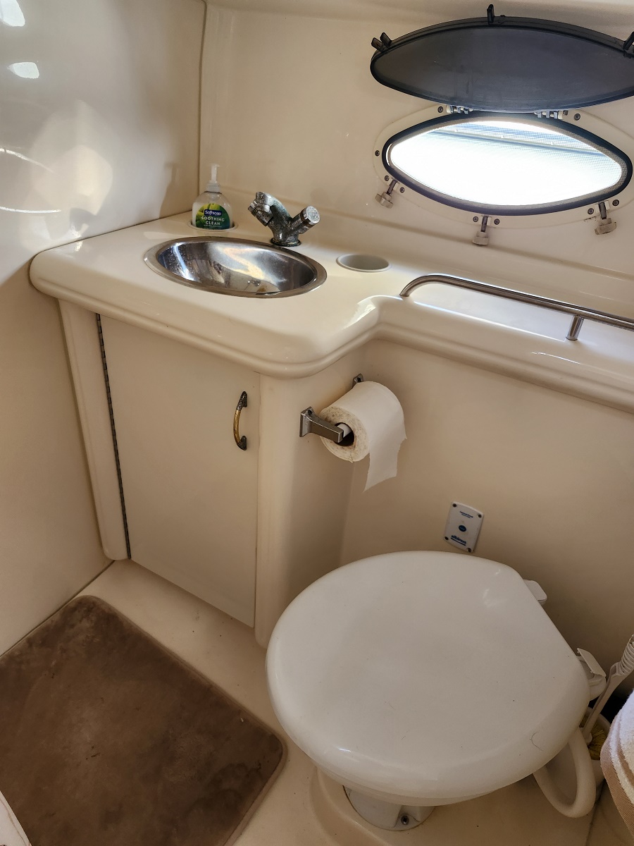 Compact boat bathroom with oval porthole window, sink, faucet, soap bottle, toilet, and tissue holder in cream cabinetry