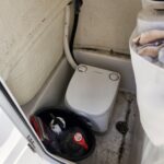 Small white portable marine toilet (Dometic) in a boat head, with black and white hoses attached and a dirty, stained floor nearby. A bucket with supplies sits next to the unit.",