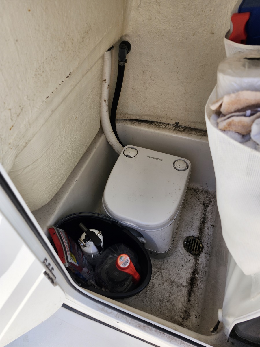 Small white portable marine toilet (Dometic) in a boat head, with black and white hoses attached and a dirty, stained floor nearby. A bucket with supplies sits next to the unit.",