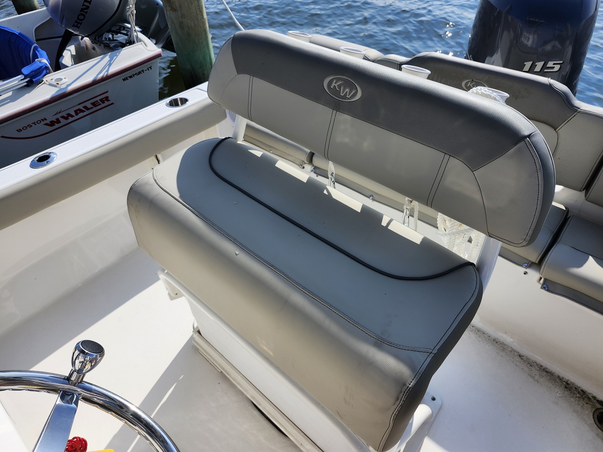 Gray boat seat with a KW logo on the backrest, mounted on a white deck with water in the background and another boat nearby by the dock